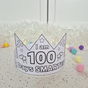 May include: A white paper crown with the text "I am 100 Days Smarter" in black. The crown has a star and heart design and is surrounded by colorful pom-poms. The crown is designed to be colored in.