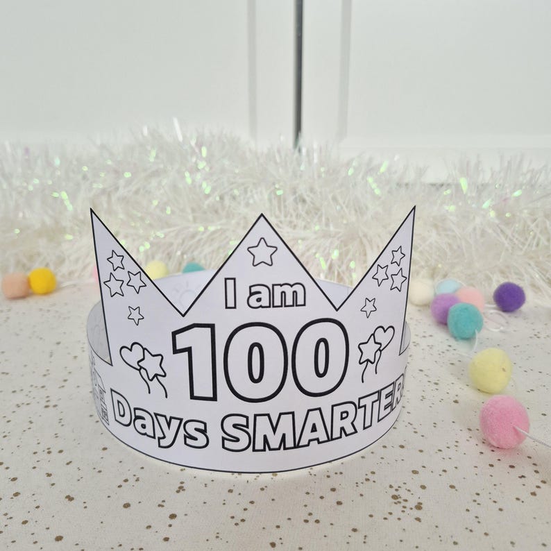 May include: A white paper crown with the text "I am 100 Days Smarter" in black. The crown has a star and heart design and is surrounded by colorful pom-poms. The crown is designed to be colored in.
