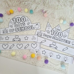 May include: Black and white paper crowns and decorative bands with the text "100 Days of School" and "I am 100 Days Smarter!" featuring school-themed illustrations. The crowns and bands are surrounded by colorful pom-poms and a white garland.