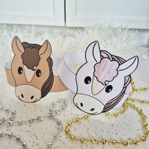 Pu&ograve; includere: Due corone di carta a tema cavallo. Una &egrave; color cuoio con criniera e ciuffo marroni, l'altra &egrave; bianca con criniera rosa e marrone. Le corone sono su una superficie bianca con collane di perline argentate e dorate.