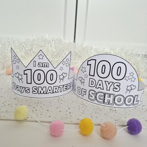 May include: Two white paper crowns with black outlines and text. One crown reads "I am 100 Days Smarter!" and the other reads "100 Days of School." Both crowns have star and flower illustrations. A garland of colorful pom-poms is in the foreground.