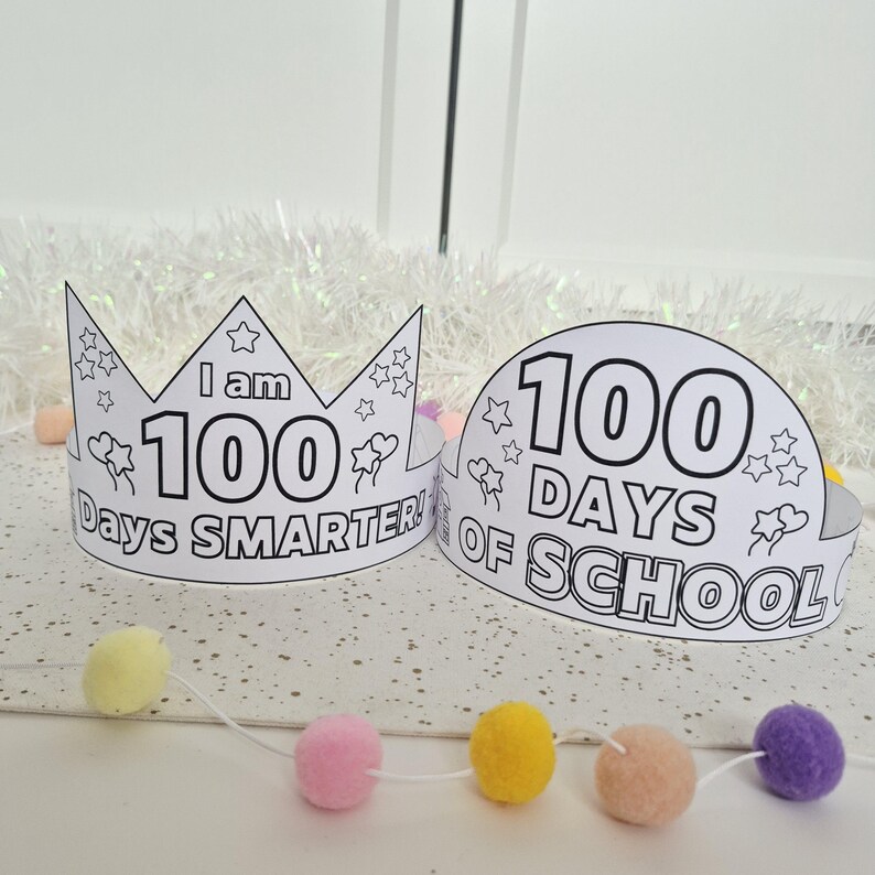 May include: Two white paper crowns with black outlines and text. One crown reads "I am 100 Days Smarter!" and the other reads "100 Days of School." Both crowns have star and flower illustrations. A garland of colorful pom-poms is in the foreground.