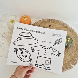 May include: A white paper sheet featuring a black and white Build a Scarecrow Craft template with a hat, head, body, and leaf outline. The text Lumong Art is printed on the page. The design includes dashed lines for cutting and glue placement areas.