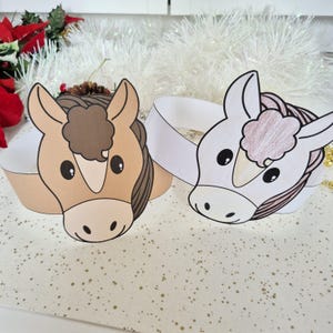 Pu&ograve; includere: Due cerchietti di carta a forma di cavallo. Uno &egrave; marrone chiaro con una criniera marrone e l'altro &egrave; bianco con una criniera rosa. I cerchietti sono su una superficie bianca con macchie dorate. Decorazioni natalizie sullo sfondo.