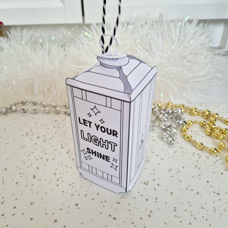 May include: A white and black paper lantern ornament with the text "LET YOUR LIGHT SHINE". The lantern has a black and white twisted cord for hanging. The ornament is decorated with star shapes.
