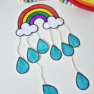 May include: A colorful rainbow-shaped craft with white clouds and teal teardrops hanging below. The rainbow features red, orange, yellow, green, and purple arches. A beaded garland and a rainbow-colored rope circle are in the background.