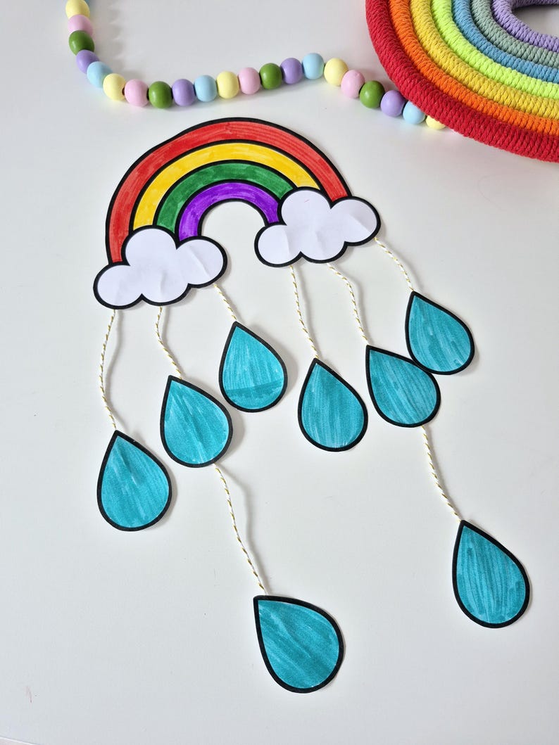 May include: A colorful rainbow-shaped craft with white clouds and teal teardrops hanging below. The rainbow features red, orange, yellow, green, and purple arches. A beaded garland and a rainbow-colored rope circle are in the background.