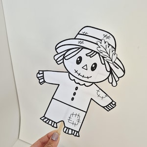 May include: A black and white line art illustration of a scarecrow puppet on a wooden stick. The scarecrow features a wide-brimmed hat with a leaf detail, a stitched mouth, round eyes, a triangular nose, and a shirt with patches on the sleeves and pants.