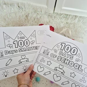 May include: Two white paper crowns with black outlines, featuring the text "I am 100 Days Smarter" and "100 Days of School." The crowns are decorated with stars, hearts, pencils, and light bulbs, perfect for a school celebration.