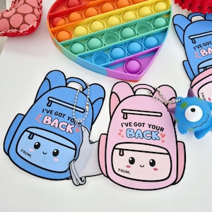 Puede incluir: Dos tarjetas coloridas con forma de mochila con el texto "Te cubro las espaldas" y una cara sonriente. Las tarjetas están unidas a un llavero con un dije de plástico transparente.