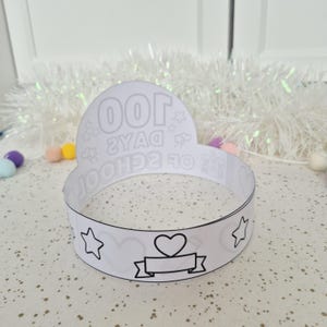 May include: A white paper crown with black outlines. The crown features the text "100 DAYS OF SCHOOL" and various illustrations, including stars, hearts, and a banner. The crown is designed for a school celebration.