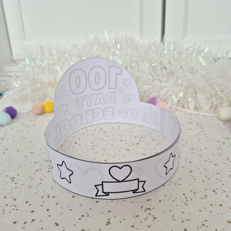 May include: A white paper crown with black outlines. The crown features the text "100 DAYS OF SCHOOL" and various illustrations, including stars, hearts, and a banner. The crown is designed for a school celebration.