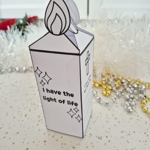 May include: A white paper candle-shaped decoration with a black outline of a flame at the top. The candle has the text "I have the light of life" and a cross design. Decorative stars are also included.