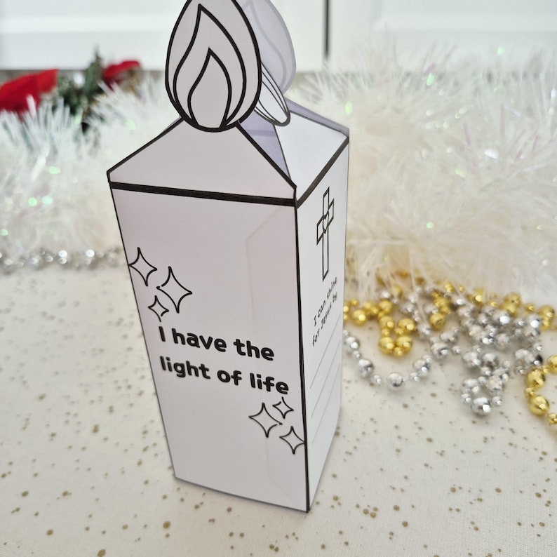 May include: A white paper candle-shaped decoration with a black outline of a flame at the top. The candle has the text "I have the light of life" and a cross design. Decorative stars are also included.