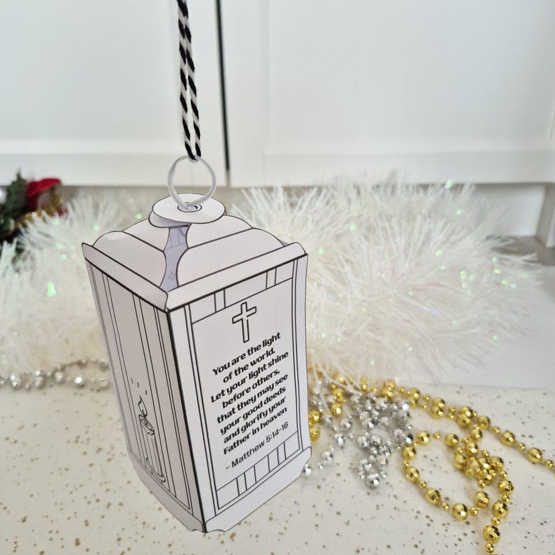May include: A white paper lantern with a black and white cord for hanging. The lantern has a cross and text from Matthew 5:14-16. The lantern is a religious-themed craft.