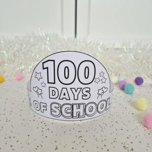 May include: A white paper crown with the text "100 DAYS OF SCHOOL" in black. The crown is decorated with stars and flowers. The background includes white tinsel and colorful pom-poms.
