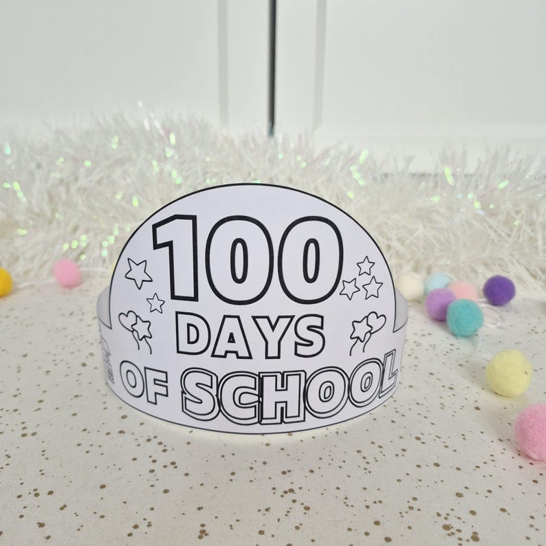 May include: A white paper crown with the text "100 DAYS OF SCHOOL" in black. The crown is decorated with stars and flowers. The background includes white tinsel and colorful pom-poms.