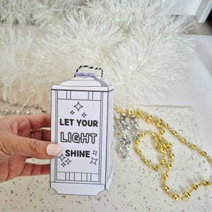 May include: A white lantern-shaped paper craft with the text "LET YOUR LIGHT SHINE" and star graphics. The lantern has a black handle and is decorated with black lines. The background includes white tinsel and gold and silver beads.