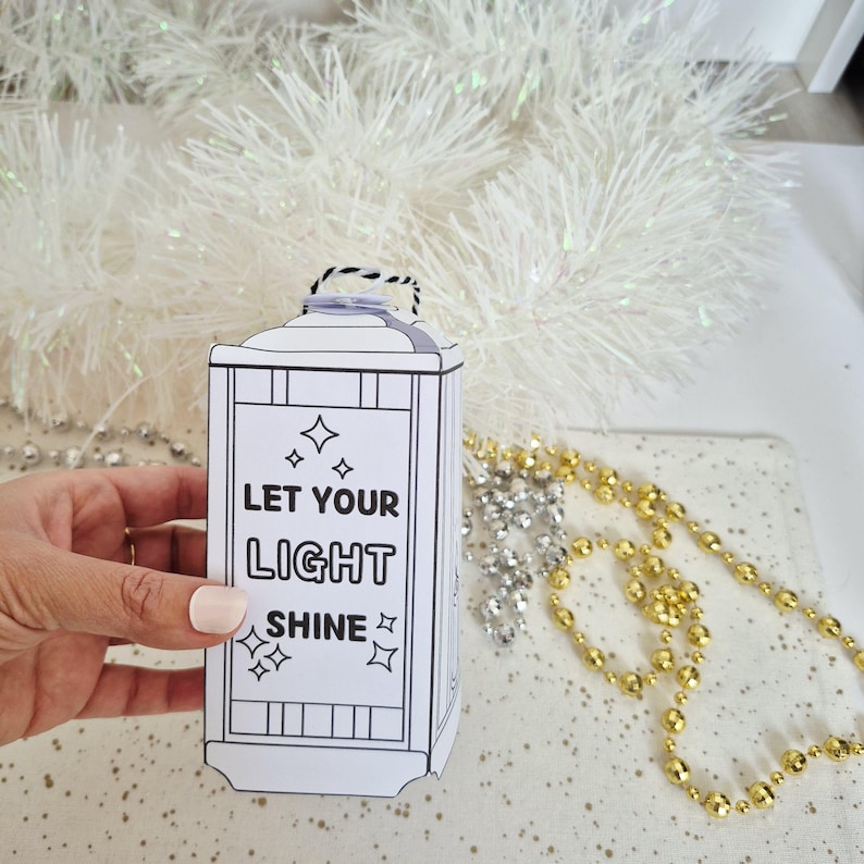 May include: A white lantern-shaped paper craft with the text "LET YOUR LIGHT SHINE" and star graphics. The lantern has a black handle and is decorated with black lines. The background includes white tinsel and gold and silver beads.