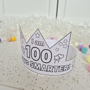May include: A white paper crown with the text "I am 100 days SMARTER!" in black. The crown is decorated with stars, balloons, and suns. Perfect for a child's 100th day of school celebration.
