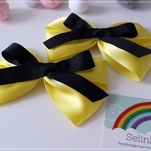 May include: Two yellow satin hair bows with black ribbon centres. The bows are handmade and are displayed on a white surface with a business card that reads "Selinish Girls handmade hair bows".