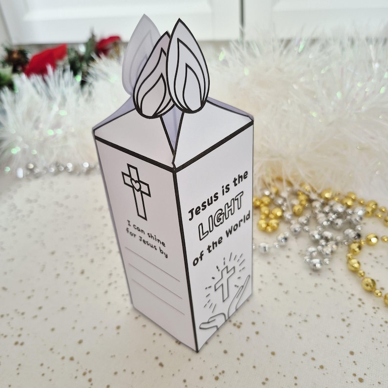 May include: A white paper candle-shaped box with black outlines and text. The box features a cross graphic and the words "Jesus is the LIGHT of the World." Two paper flames are attached to the top. The box is decorated with gold and silver beads.
