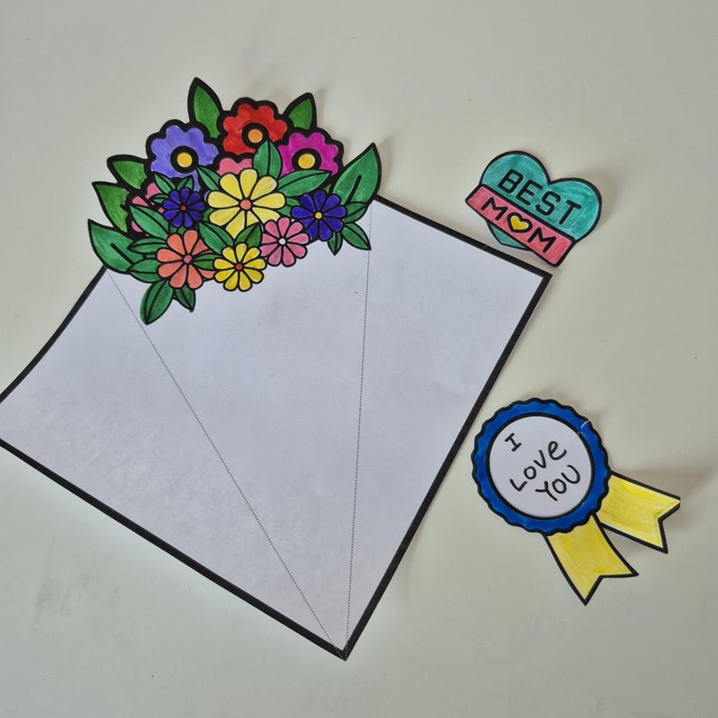 May include: A handmade Mother's Day card with a bouquet of colorful flowers, a heart-shaped badge that says "Best Mom", and a blue ribbon that says "I Love You". The card is white with a black border.