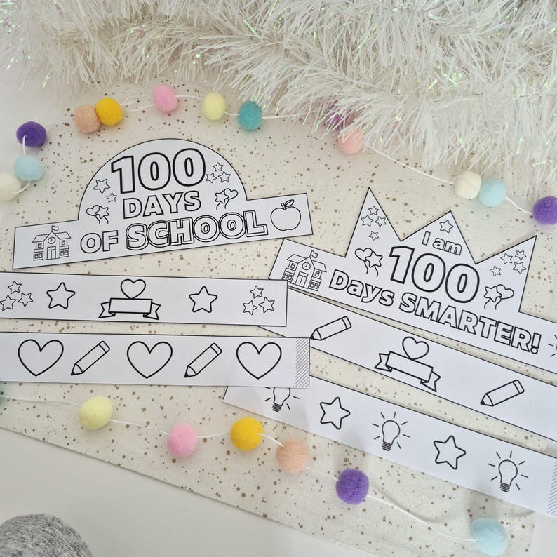 May include: White paper crowns and decorative bands with the text "100 Days of School" and "I am 100 Days Smarter!" are displayed. The crowns and bands feature outlines of stars, hearts, pencils, and lightbulbs. A garland of pastel pom-poms and white tinsel are in the background.