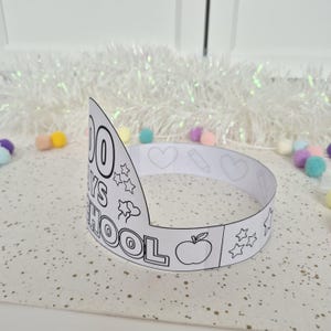 May include: A white paper crown with the text "100 DAYS OF SCHOOL" and various outline drawings, including stars, hearts, a pencil, and an apple. The crown is designed for coloring.