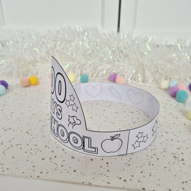 May include: A white paper crown with the text "100 DAYS OF SCHOOL" and various outline drawings, including stars, hearts, a pencil, and an apple. The crown is designed for coloring.