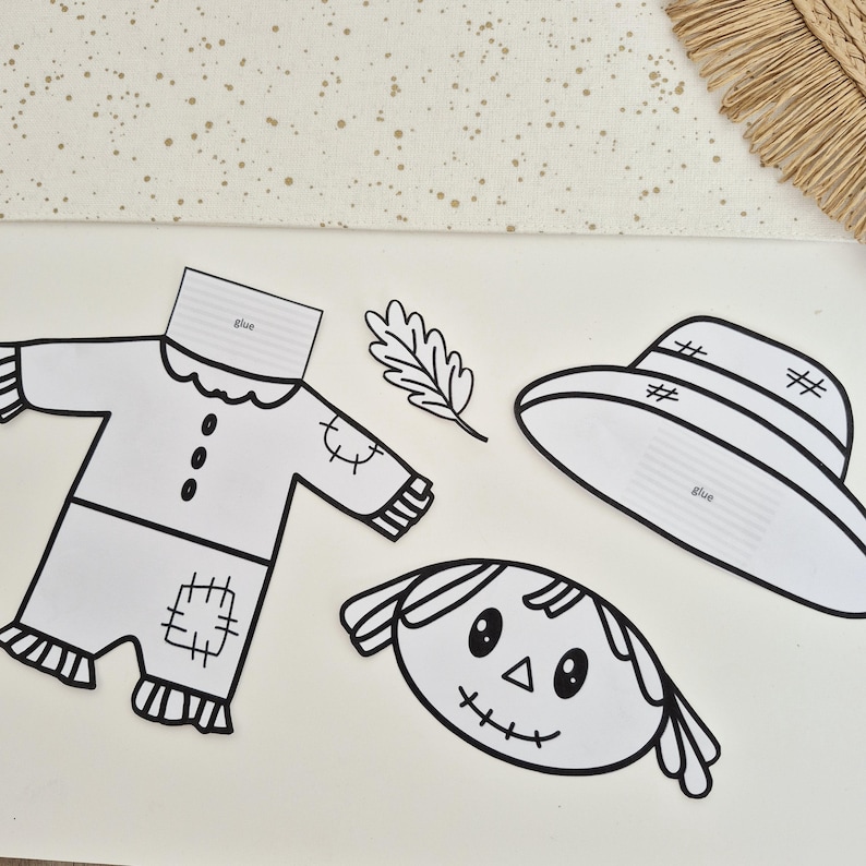 May include: A black and white paper craft set featuring a scarecrow outfit, a hat, a leaf, and a face. The clothing and hat pieces include a tab labeled glue for assembly. The scarecrow face has stitched eyes, a triangular nose, and a stitched mouth.