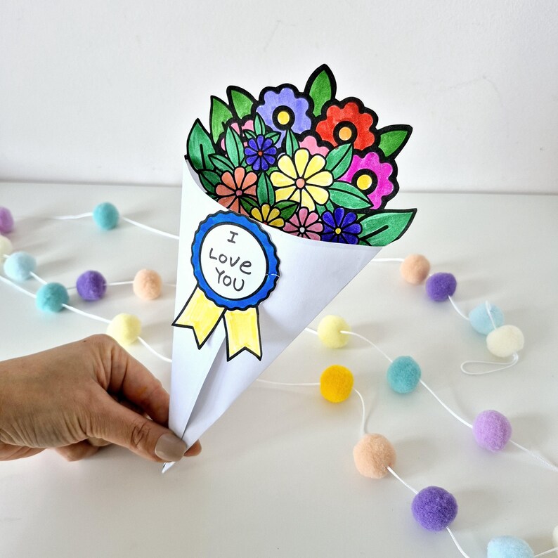 May include: A handmade paper bouquet with colorful flowers, including red, purple, yellow, and pink blooms, is held in a white cone. The bouquet has a blue and yellow ribbon with the text "I Love You". A string of pastel pom-poms decorates the background.