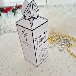 May include: A white paper candle-shaped box with black line art. The box features a cross, the text "Jesus is the LIGHT of the World", and space for writing. Two flame cutouts are at the top. Religious themed craft.