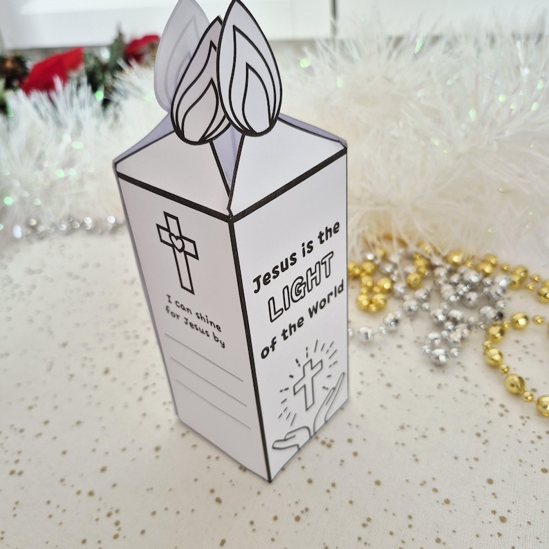 May include: A white paper candle-shaped box with black line art. The box features a cross, the text "Jesus is the LIGHT of the World", and space for writing. Two flame cutouts are at the top. Religious themed craft.