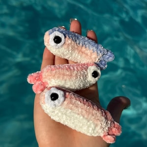 以下が含まれることがあります： 柔らかくふわふわした質感の、かぎ針編みの魚の形をした小さなぬいぐるみ3つ。それぞれのぬいぐるみは、白い腹部、ピンクまたは青のグラデーションの背中、そして黒いプラスチック製の安全な目が1つ付いています。魚は縦に積み重なっています。