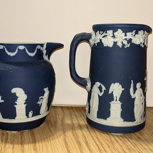 May include: Two blue and white ceramic pitchers with white designs. The pitcher on the left is rounder and has a white design of figures and a garland. The pitcher on the right is taller and has a white design of figures and grape vines.