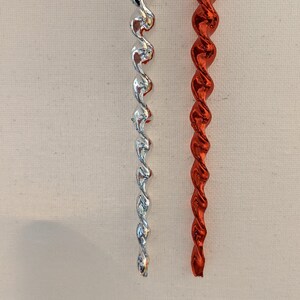 May include: Two decorative Christmas ornaments. One is silver, the other is red. Both have a twisted, icicle-like design and are suspended by thin strings. The ornaments are against a neutral background.
