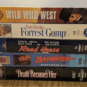 May include: A stack of five VHS tapes, including "Wild Wild West", "Forrest Gump", "Road House", "Baywatch: Nightmare Bay", and "Death Becomes Her".