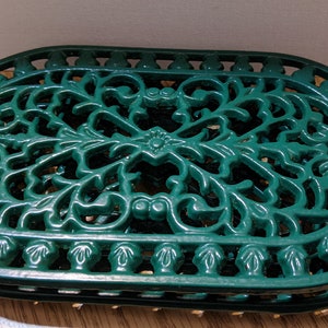 May include: A green cast iron trivet with an oval shape and a decorative floral design. The trivet is made of metal and has a glossy finish.