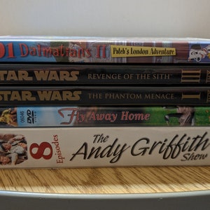 May include: A stack of five DVDs. Titles include "101 Dalmatians II", "Star Wars: Revenge of the Sith", "Star Wars: The Phantom Menace", "Fly Away Home", and "The Andy Griffith Show". The DVDs are stacked on a wooden surface.