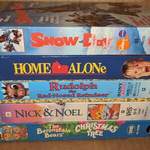 May include: A stack of six VHS tapes featuring classic holiday movies, including "Snow Day", "Home Alone", "Rudolph the Red-Nosed Reindeer", "Nick & Noel", and "The Berenstain Bears' Christmas Tree".