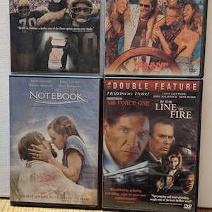 May include: Four DVD cases with different movie titles. The top left case is for the movie "Rudy" and features a football player in a white and blue uniform. The top right case is for the movie "Captain Ron" and features a group of people on a boat. The bottom left case is for the movie "The Notebook" and features a couple kissing in the rain. The bottom right case is for the movie "Air Force One" and features Harrison Ford and Clint Eastwood.