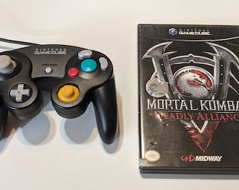 May include: A black Nintendo GameCube controller and a black game case for Mortal Kombat: Deadly Alliance. The game case has a red and black dragon design with the game title in white.