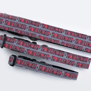 May include: Three gray dog collars with black buckles. The collars are printed with the repeated text "Troublemaker" in pink.