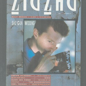 May include: A magazine cover featuring a man with short brown hair, wearing a blue shirt, looking down at a magazine. The magazine cover is for Zig Zag, a monthly music magazine, and features the text "Sho'Gun Wedding" and "Billy Bragg / In Japan / In His Own Words".