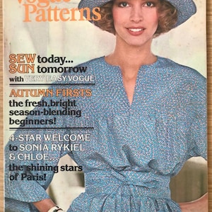 May include: A vintage Vogue Patterns magazine cover featuring a woman wearing a blue and white patterned dress and a wide-brimmed hat. The cover text reads "Sew today... Sun tomorrow with Very Easy Vogue" and "Autumn Firsts".