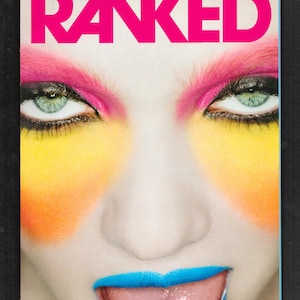Peut inclure: Une couverture de magazine avec le mot "RANKED" en grandes lettres roses. La couverture présente un gros plan d'un visage avec un maquillage éclatant, notamment un fard à paupières rose, un blush jaune et orange et un rouge à lèvres bleu. La personne sort sa langue.