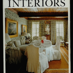 May include: A magazine cover for "The World of Interiors" magazine, featuring a photo of a dining room with a white tablecloth, a striped sofa, and a wooden ceiling.