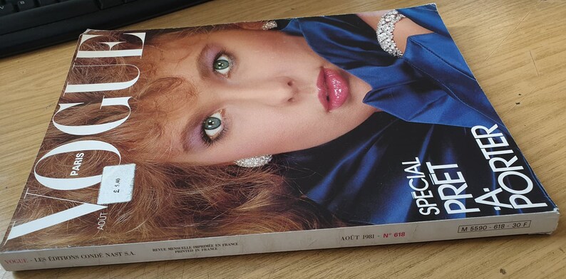 May include: A vintage Vogue Paris magazine cover from August 1981, featuring a woman with long red hair wearing a blue satin top and a diamond necklace. The cover text reads "Special Pret-a-Porter".