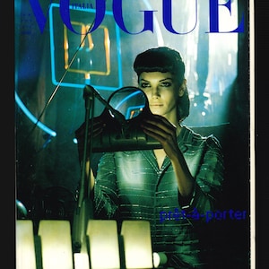 May include: A Vogue Italia magazine cover featuring a woman in a green and silver patterned dress holding a silver handbag. The cover text reads "prêt-à-porter" and "PRADA".